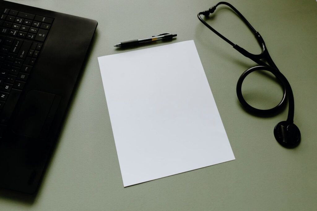 A laptop, pen, paper and stethoscope on a desk