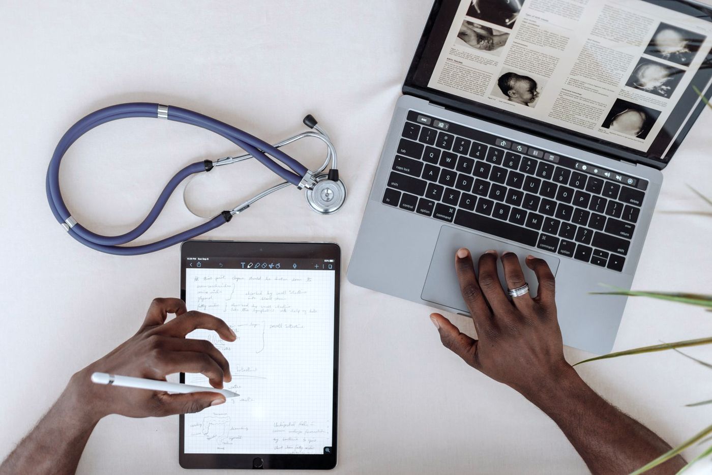 Hands using a tablet and laptop beside a stethoscope