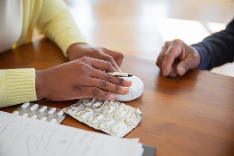 Two people talking through medicines at a table, with notes and blister packs